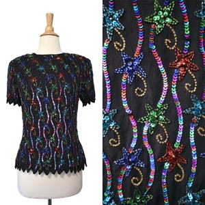 May include: Black sequined top with short sleeves. The top is adorned with colorful star-shaped sequins and wavy lines of sequins in various colors. The neckline and hemline have a scalloped edge. The top is displayed on a mannequin.