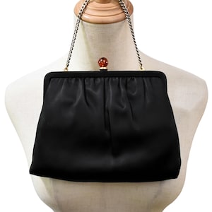 Può includere: Pochette in raso nero con tracolla a catena argentata. La chiusura presenta una pietra rotonda color ambra. La borsa è esposta su un manichino. Una classica borsa da sera.