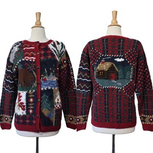 May include: A long-sleeve knit cardigan featuring a patchwork design with red, navy, and green colors. The front displays a bear and forest scene, while the back features a cabin in the woods. The garment has a button-front closure and ribbed trim.