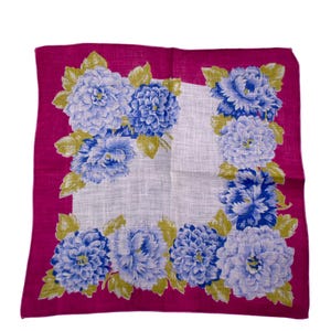 Puede incluir: Un pañuelo cuadrado con estampado floral, con un borde magenta y un centro blanco. El centro presenta flores azules y celeste con hojas verdes. El pañuelo está hecho de tela ligera.