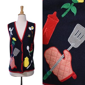 May include: A navy blue sleeveless knit vest featuring a V-neckline with red trim and button closure. The vest is decorated with various embroidered kitchen-themed appliqués including a spatula, a red and white checkered oven mitt, and food items.