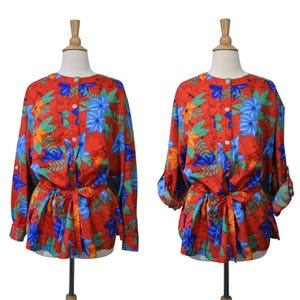 May include: A red floral blouse with long sleeves and a tie waist. The blouse features a vibrant pattern of blue, green, and yellow flowers and leaves. The sleeves can be rolled up and buttoned. The blouse is displayed on a mannequin.