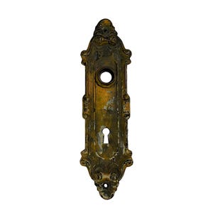 Op de afbeelding: Oud, sierlijk deurbeslag met een sleutelgat en een rond gat erboven. De plaat heeft een doffe messingkleur met decoratieve krullen en een rechthoekige vorm. Het metaal vertoont tekenen van ouderdom en slijtage.