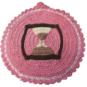 May include: A round, pink crocheted doily with a brown hourglass design in the center. The doily has a scalloped edge with white and pink trim. The hourglass is filled with beige and brown yarn.