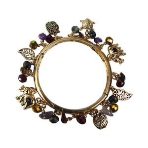 May include: A gold-toned bangle bracelet adorned with various charms and colorful beads. The charms include animal figures, leaves, and other decorative elements. The bracelet is set against a white background.