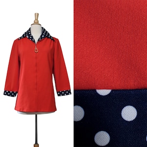 May include: A red zip-up jacket with a navy blue and white polka dot collar and cuffs. The jacket has a gold-colored zipper pull. The fabric appears to be a soft, textured material. The jacket is displayed on a mannequin.