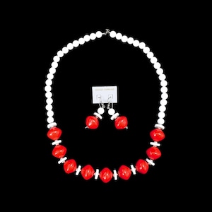 Puede incluir: Un conjunto de collar y pendientes con una cadena de cuentas blancas intercaladas con cuentas rojas cuadradas. Los pendientes y el collar se muestran sobre un fondo negro.