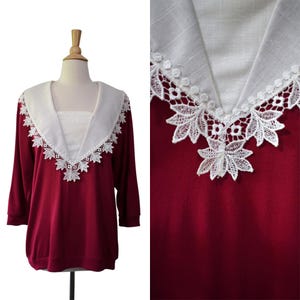 May include: A burgundy long-sleeve top with a white collar and intricate white lace detailing. The collar is a wide V-neck shape, and the lace forms a decorative border along the neckline. The top is displayed on a mannequin.