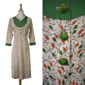 May include: A knee-length dress featuring a cream base with a repeating pattern of small orange and green lantern-like shapes on thin black branches. The dress has a solid green collar, green sleeve cuffs, and three green buttons.