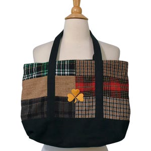 May include: A tote bag featuring a patchwork design of various plaid and tweed fabrics in green, brown, red, and black. The bag has solid black fabric handles and a solid black base. A bright yellow embroidered shamrock is centered on the front.