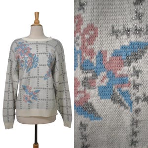 May include: A light gray sweater with a floral design in pink and blue. The sweater has a grid pattern and a cream-colored neckline and hem. The long-sleeved sweater is displayed on a mannequin.