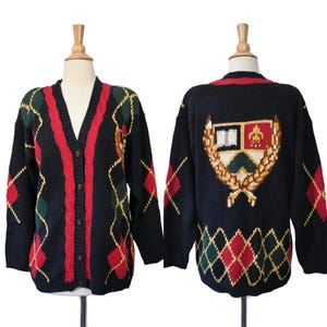 May include: A black knit cardigan featuring a V-neckline with red cable-knit trim and a button-front closure. The garment displays a multi-coloured argyle pattern in red, green, and yellow, with a large crest emblem on the back featuring a shield.