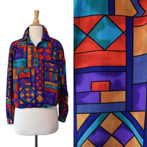 Puede incluir: Una blusa de manga larga con un vibrante patrón geométrico en tonos morado, naranja, azul verdoso y rojo. La camisa tiene cuello, una tapeta frontal con botones parciales y un dobladillo elástico en la cintura para un ajuste ceñido.