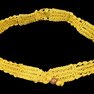 May include: A handmade yellow macrame belt with a decorative pattern. The belt features a small, round, brown bead at the center. The background is solid black.