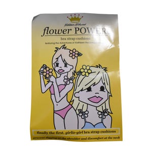 May include: A yellow product package for Kathleen Kirkwood flower POWER bra strap cushions. The packaging features a cartoon illustration of a blonde character with flower hair accessories, wearing pink undergarments and blue straps.