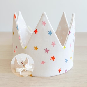 Könnte beinhalten: Eine weiße Stoffkrone mit einem bunten Sternenmuster. Die Krone ist aus Filz und hat eine spitze Spitze. Sie ist perfekt für eine Geburtstagsfeier oder einen anderen besonderen Anlass.