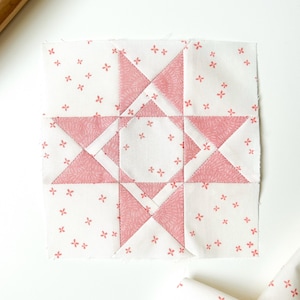 May include: A square quilt block featuring a star design in shades of pink and white. The star is composed of triangles and squares, with a floral pattern on the white background. The block is likely for a larger quilt project.
