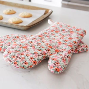 May include: A pair of floral oven gloves with a white background and a pink and orange floral pattern. The gloves are quilted and have a loop for hanging.