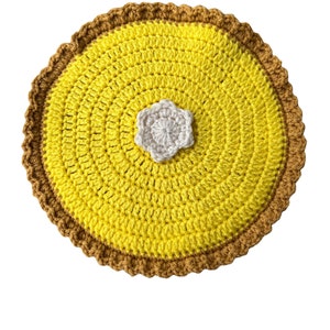 May include: A handmade, crocheted pie-shaped item with a yellow filling and a brown crust. A small, white, flower-shaped embellishment is centered on the pie. The item is likely a decorative piece or a coaster.