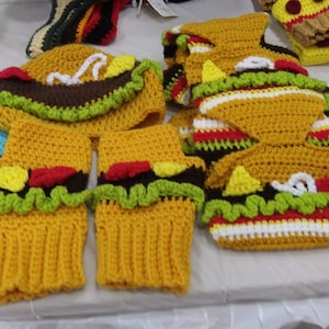 May include: A collection of handmade crochet food-themed items, including hats and hand warmers. The items are designed to resemble a hamburger and a taco, with yellow, brown, red, green, and white yarn.