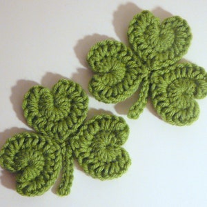 Può includere: Due quadrifogli in crochet verde. I quadrifogli sono realizzati con un filo verde chiaro e hanno una superficie testurizzata e irregolare.
