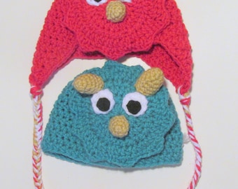 May include: Two crocheted hats with dinosaur faces. One hat is red with white eyes and a yellow nose. The other hat is teal with white eyes and a yellow nose. Both hats have long, braided ties with red, white, and yellow stripes.