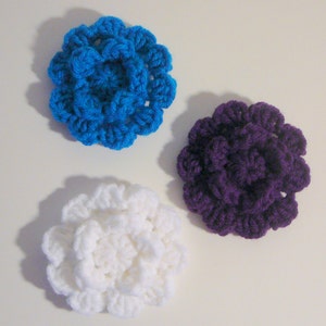May include: Three crocheted flowers in different colours: white, purple, and blue. The flowers are made with a textured stitch and have a layered appearance.