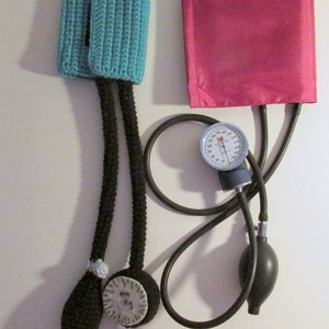 Peut inclure: Un brassard de tension art&eacute;rielle en crochet noir et turquoise avec une poire noire et un manom&egrave;tre argent&eacute;. Le manom&egrave;tre a une aiguille noire et des marques blanches.