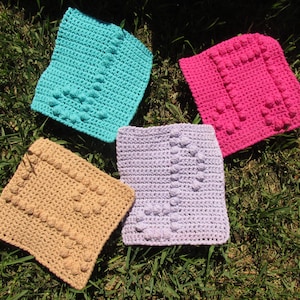 May include: Four crocheted dishcloths in different colors: teal, beige, lavender, and pink. Each dishcloth has a textured design with raised bumps.