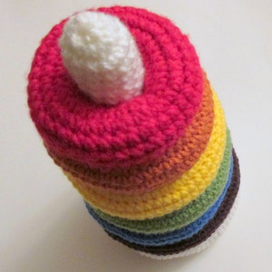 Puede incluir: Una pila de anillos de crochet arcoíris con una parte superior de crochet blanco. Los anillos son de color rojo, naranja, amarillo, verde, azul y morado.