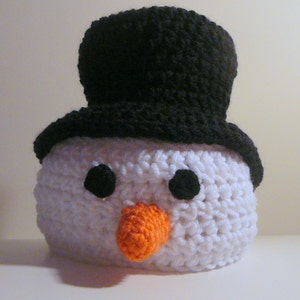 May include: White crocheted snowman head with black eyes, orange carrot nose, and a black crocheted top hat.