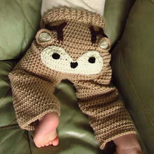 May include: Brown crocheted baby trousers with a deer face design. The deer face has brown antlers, black eyes, and a white nose.