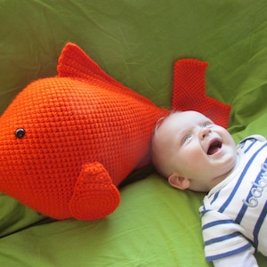 May include: A crocheted orange fish toy with a black button eye, lying on a green blanket next to a smiling baby wearing a striped shirt with the text "baby girl" embroidered on it.