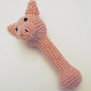 May include: A pink crocheted pig rattle toy with black eyes. The rattle is shaped like a pig with a long handle.