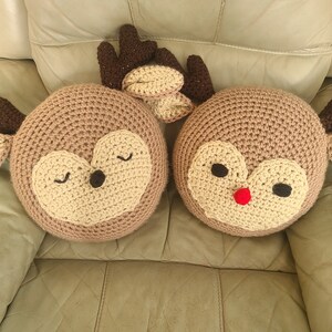 May include: Two crocheted reindeer pillows, one with a red nose and the other with closed eyes. Both pillows are brown with beige accents.