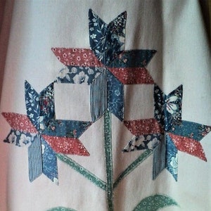 Puede incluir: Una tela blanca con un diseño floral azul, rojo y verde. Las flores están hechas de tela de patchwork y tienen forma de estrella.