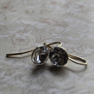 May include: A pair of gold-coloured drop earrings featuring clear, faceted crystal stones. The earrings have a simple design, with the crystals reflecting light. The background is a soft, patterned surface.