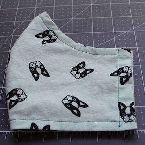 May include: A light blue fabric with a repeating pattern of black French Bulldog silhouettes. The fabric is sewn into a mask shape with a green seam around the edges.