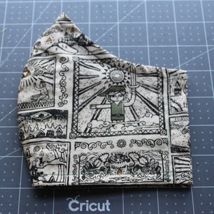 May include: A beige and black face mask featuring a repeating pattern of a video game character holding a sword, sun, and other graphic elements. The mask is made of fabric and has a curved shape to fit the face.