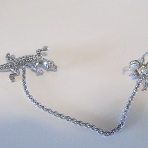Könnte beinhalten: Silberfarbene Brosche mit einem Eidechsen- und Spinnen-Motiv, verbunden durch eine Kette. Beide Figuren sind mit kleinen, glitzernden Kristallen besetzt. Die Eidechse befindet sich links, die Spinne rechts.