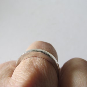 May include: A silver ring with a smooth, curved design. The ring is shown on a finger, with a focus on the polished metal band. The ring's simple, elegant style suggests it could be a wedding band or a minimalist fashion accessory.