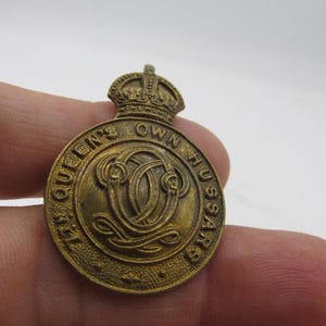 Puede incluir: Un emblema circular de lat&oacute;n con el texto "THE QUEEN'S OWN HUSSARS" que rodea un dise&ntilde;o central. El emblema tiene una corona en la parte superior. El color general es un dorado deslustrado. El emblema se sostiene en una mano.