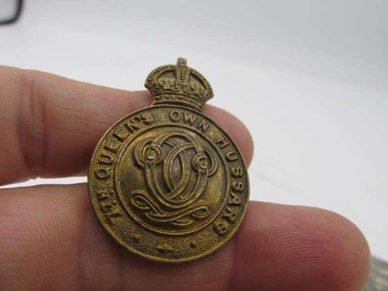 Puede incluir: Un emblema circular de lat&oacute;n con el texto "THE QUEEN'S OWN HUSSARS" que rodea un dise&ntilde;o central. El emblema tiene una corona en la parte superior. El color general es un dorado deslustrado. El emblema se sostiene en una mano.