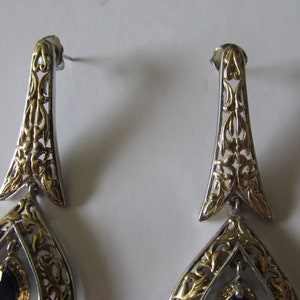 Op de afbeelding: Een paar zilveren en gouden oorhangers met een zwarte edelsteen. De oorbellen hebben een filigraanontwerp met ingewikkelde details.