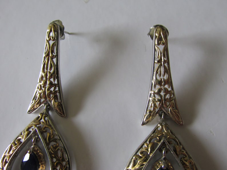 Op de afbeelding: Een paar zilveren en gouden oorhangers met een zwarte edelsteen. De oorbellen hebben een filigraanontwerp met ingewikkelde details.