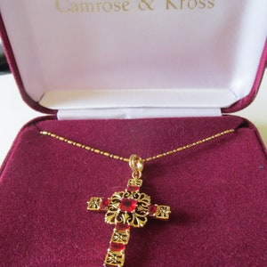 May include: A gold-toned cross pendant necklace with red gemstones, displayed in a burgundy jewelry box. The cross features intricate filigree detailing and rectangular red stones. The box is lined with burgundy velvet and has the text "Camrose & Kross" on the inside.