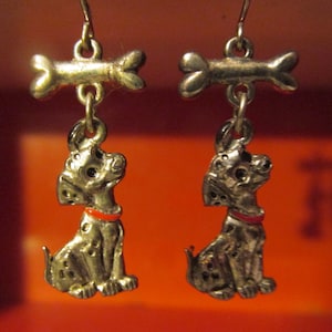May include: A pair of silver-toned dog earrings. Each earring features a sitting Dalmatian dog charm with black spots and a red collar, suspended from a bone-shaped top. The earrings are designed with a hook closure.