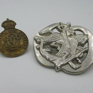 Puede incluir: Dos insignias militares vintage. Una es una insignia circular de lat&oacute;n con una corona y el texto "THE QUEEN'S OWN HUSSARS". La otra es una insignia de color plateado con un dise&ntilde;o detallado de un caballo y un jinete.