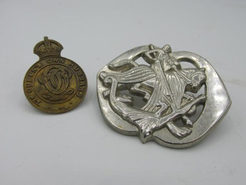 Puede incluir: Dos insignias militares vintage. Una es una insignia circular de lat&oacute;n con una corona y el texto "THE QUEEN'S OWN HUSSARS". La otra es una insignia de color plateado con un dise&ntilde;o detallado de un caballo y un jinete.