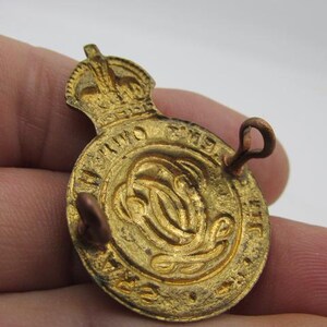 Puede incluir: Una insignia met&aacute;lica dorada, de forma circular, con una corona en la parte superior. La insignia presenta texto en relieve alrededor del borde y un dise&ntilde;o central. Dos presillas est&aacute;n unidas a la parte posterior para asegurar la insignia.